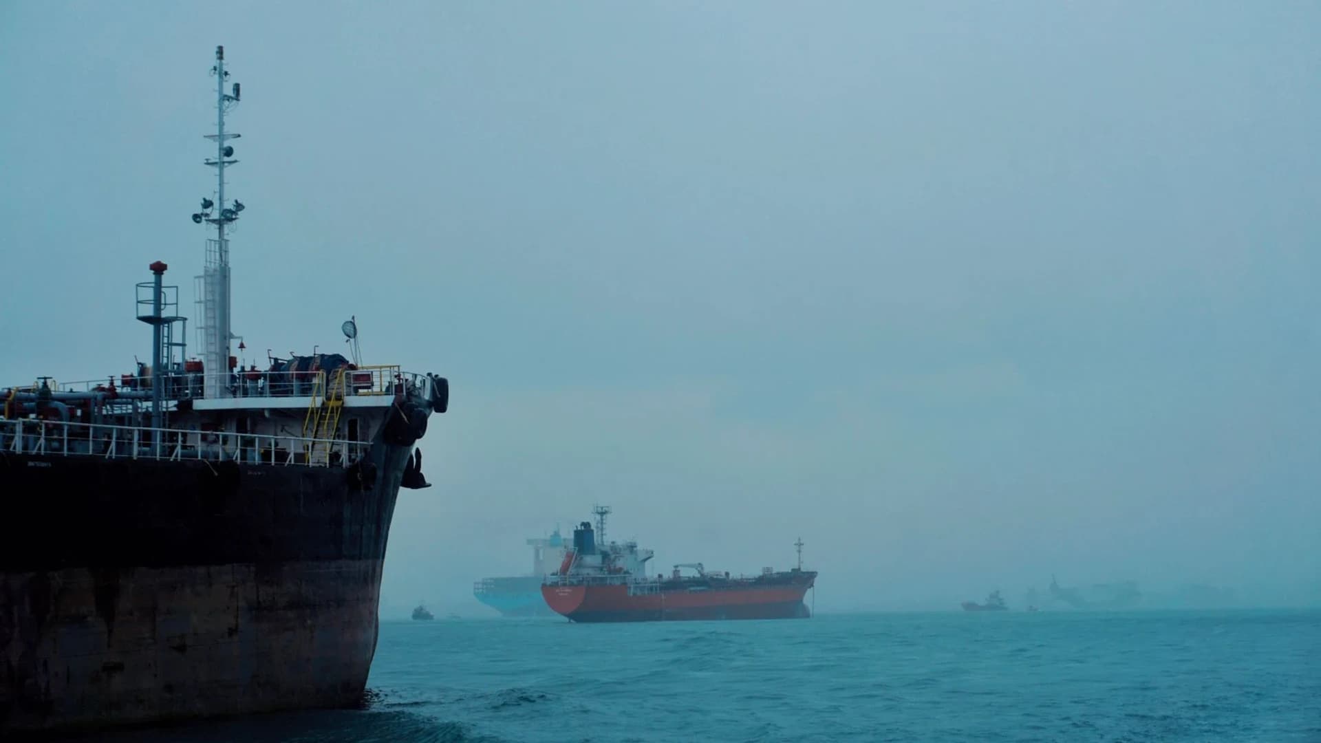 A fleet of cargo ships docked near oil storage tanks along a serene coastline with a clear blue sky above.